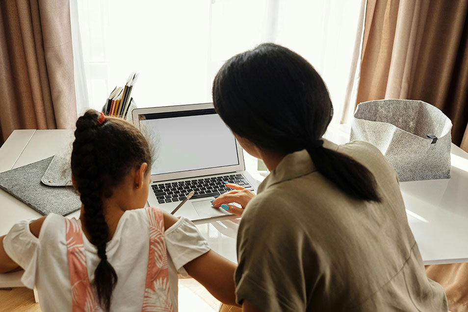 Your_Young_Business_Needs_Northout Image of woman at laptop with child by her side seated at a table, the child writing notes as the woman uses the touchpad.