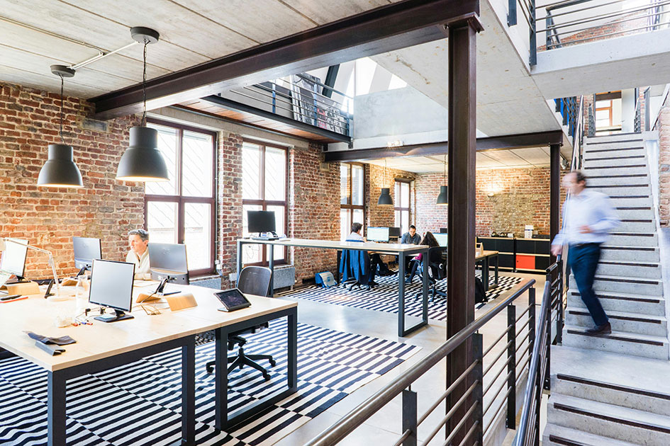 Ppen plan office with open plan stairway leading to atrium. Man walking down stairway, desks with monitors, a few people sitting at desks further away and a man seated working at desk in foreground.
