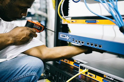 Photograph of man in white t-shirt and jeans holding a small power tool/electric screw driver in his right hand and lifting up a switch box with his left hand. The switchbox he is holding is within a server cage which contains other switch boxes and cables between them. He is working on the network of a business.