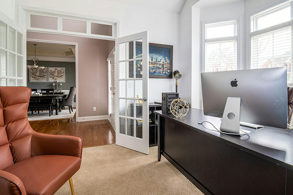 Photograph of home office with iMac on the desk and a mid-century modern leather chair opposite the desk, the office doors are glass panelled white painted french doors, opening to a hallwall through which you can see a dining room table and chairs.