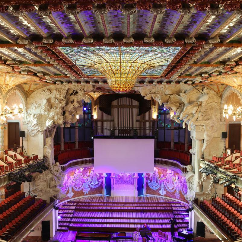 Town_Hall_01 Photograph of large town hall with chairs on two tiered system and large central ornate glass panel in the ceiling surrounded by plaster sconces held by pillars with cargins of figures and horses. A modern white drop screen dangles infront of a large music organ against the back wall/stage area.