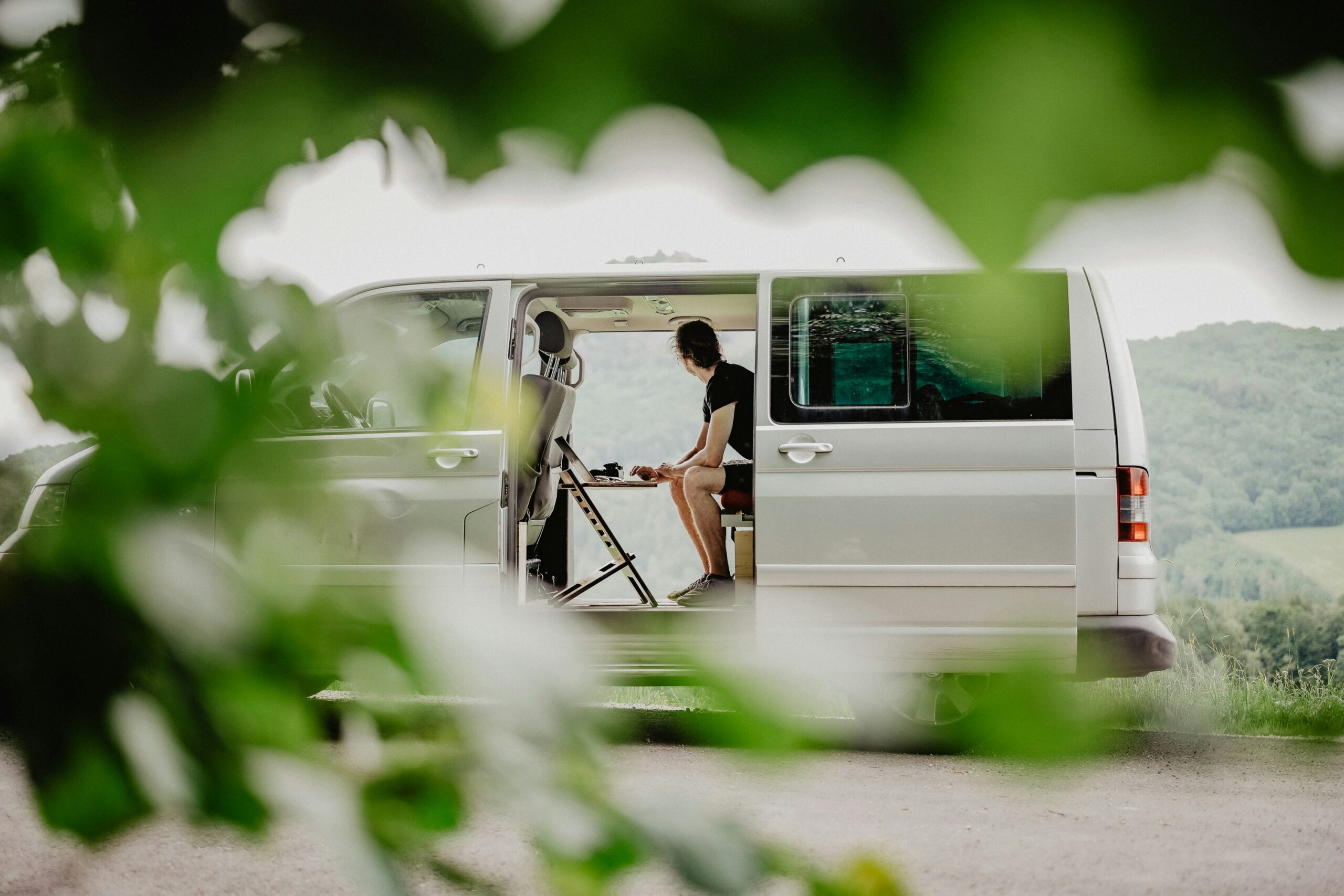 Photograph of campervan with double side doors both open so that you can see the countryside through it. Inside the campervan a man is seated at a wooden folding desk with laptop looking out over the view, his hand poised at the laptop.